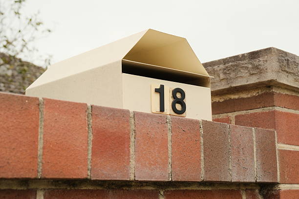 Beige metal mailbox with the number 18 on a brick wall. Overcast sky above.
