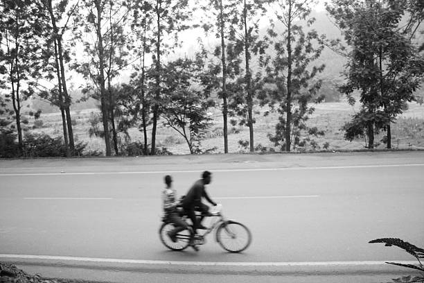 An evocative color photograph captured with a panning technique, creating sharp focus on the central subject and dramatic motion blur in the background. A single motorcycle rider, wearing a red shirt and helmet, speeds along a wide, rural highway. The surrounding landscape, including trees and fields, is streaked with movement, emphasizing speed, travel, commuting, and the dynamic rhythm of life outside the city.