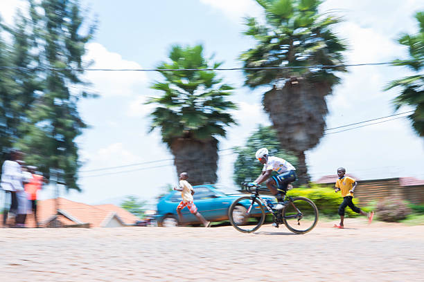 A high-energy color photograph captured using a panning technique, resulting in heavy motion blur. The central focus is a cyclist wearing a helmet and specialized cycling gear on a road bike, captured mid-speed. The background is a mix of urban and tropical elements: tall palm trees, a moving blue car, a red-tiled roof, and local children running alongside, emphasizing the excitement and speed of the race or training. This image is excellent for themes of competitive sport, fitness, activity, and dynamic urban life.