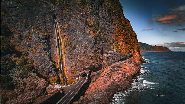 Aerial view of a scenic coastal road and tunnel carved into steep cliffs on Madeira Island, Portugal, with a slender waterfall cascading down the rock face towards the Atlantic Ocean at sunset.