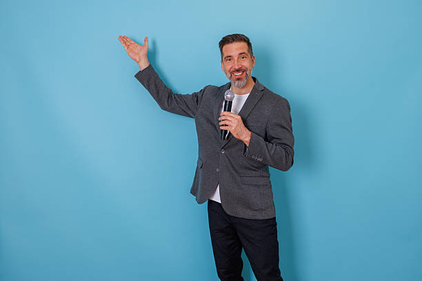 Mid adult man smiling, holding a microphone, and presenting while gesturing with an outstretched arm, indicating clear direction