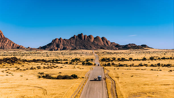 A lone vehicle travels a straight gravel road across golden Namibian plains toward the dramatic Spitzkoppe rock formations under a clear blue sky, conveying adventure and remote wilderness.