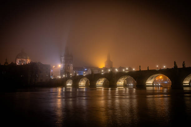 Long exposure view of Charles Bridge in Prague during a foggy winter night with glowing arches and Old Town lights reflecting in the Vltava River.