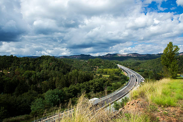 A multi-lane highway curves over a bridge through a forested valley, with trucks and cars under a dramatic cloudy sky.