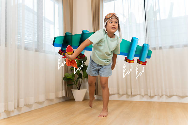 Creative child wearing aviator hat pretending to fly with a toy rocket and illustrated wings at home, showing imagination, creativity, playful learning, childhood dreams, and joyful indoor lifestyle.