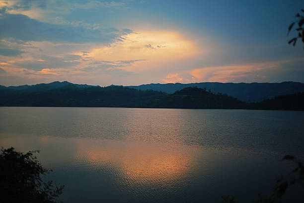 A peaceful lake at sunset with soft clouds glowing in warm tones and their reflections shimmering on the water. Dark mountain silhouettes frame the horizon, creating a calm and scenic natural landscape ideal for travel, nature, relaxation, and outdoor themes.