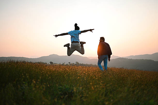 Two friends enjoy a carefree moment at sunrise, one jumping mid-air while the other watches, set against rolling hills and a grassy mountain meadow. Warm golden light, open landscape, and natural surroundings convey friendship, freedom, adventure, travel, and outdoor lifestyle themes.