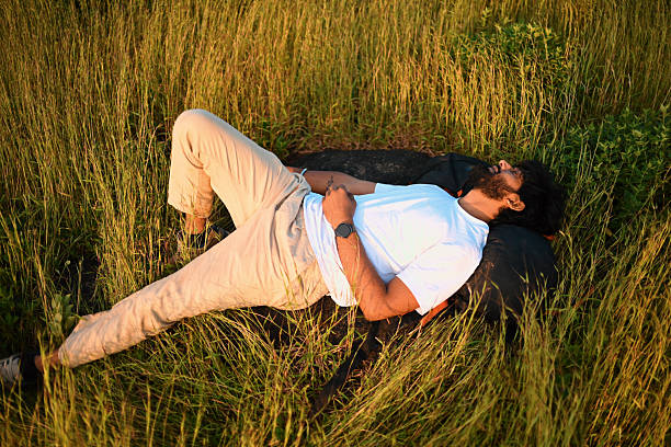 Young adult man lying on his back in a grassy meadow, resting on a backpack and enjoying a quiet moment outdoors during golden hour. Casual clothing, relaxed posture, and natural surroundings convey themes of relaxation, mindfulness, travel, freedom, and connection with nature. Horizontal, no visible logos.