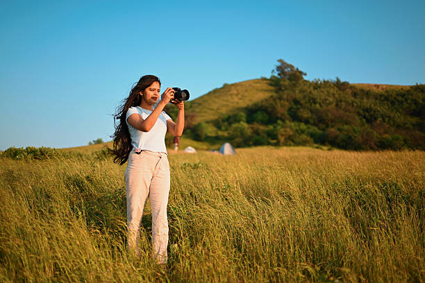 Young adult woman standing in a grassy meadow, taking photographs with a digital camera while overlooking a layered mountain landscape at sunrise. Soft golden light and open scenery convey themes of photography, travel, creativity, exploration, freedom, and connection with nature. Horizontal composition with copy space, no visible logos.