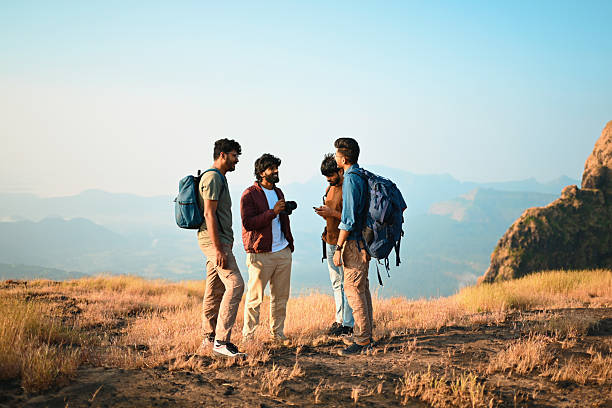Four young men with backpacks pause on a grassy hilltop while checking directions on a smartphone, surrounded by panoramic mountain landscapes. The image captures themes of adventure travel, trekking, friendship, teamwork, outdoor exploration, navigation, and modern travel planning in nature. Ideal for concepts related to hiking, youth travel, digital navigation, weekend getaways, and group bonding outdoors.