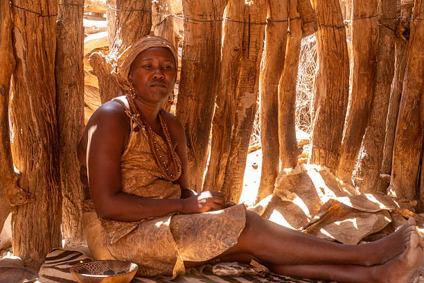 Namibia, Damaraland, Daures - Oktober 31, 2025: A woman sits quietly on the ground in a wooden shelter. She wears simple clothing and jewelry while looking thoughtfully into the distance as sunlight filters through the wooden beams.