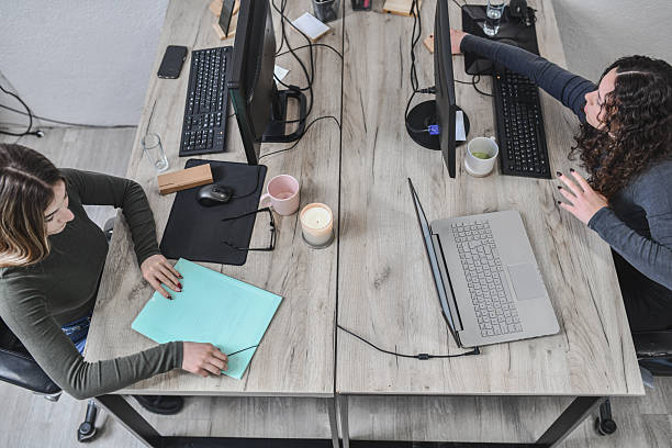 istock Two Women Working at Shared Desk with Computers, Documents, and Coffee in Modern Office 2252230085