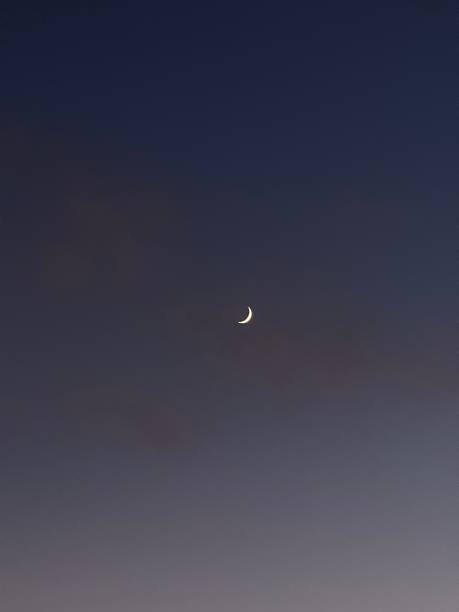 Thin crescent moon visible in a smooth twilight sky, minimal composition with soft gradients of blue and gray, representing nightfall and celestial beauty.