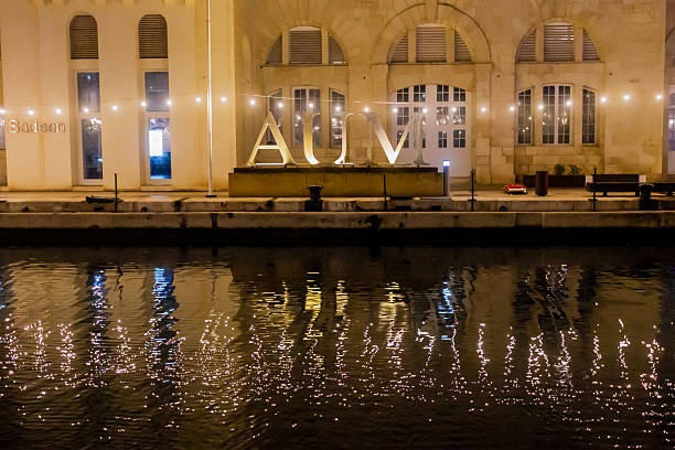 Cospicua, Malta - December 7th, 2025, Evening Waterfront With Illuminated AUM Letters Reflecting in Calm Canal Beside Historic Building