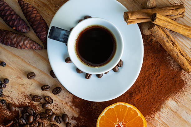 Cup of black coffee surrounded by scattered beans, cinnamon sticks, orange slice, and pine cones on wooden table