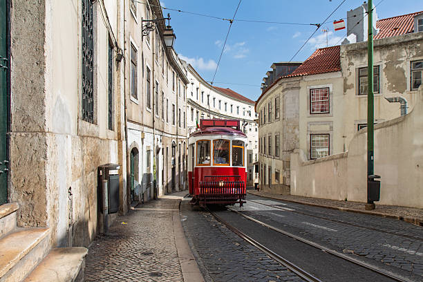 A red tourist tram traveling through the narrow, winding cobblestone streets of Lisbon's historic district on a sunny day in Portugal.