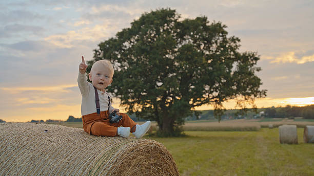 Young child perched on round hay bale in rural agricultural field during golden hour lighting, gesturing with raised hand toward distant landscape and large tree.