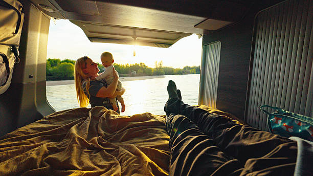 Mother in camper van holding baby boy, enjoying the view of a forested river through the open door. Van interior provides a comfortable setting for relaxation during the day.