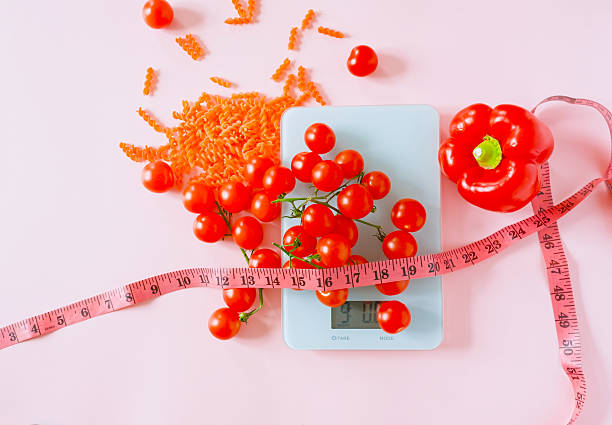 Minimal flat lay of fresh tomatoes, red bell pepper, and pasta arranged on a digital kitchen scale with a measuring tape on a pastel pink background. The image visualizes healthy eating, portion control, and illness prevention through balanced nutrition. Clean composition with generous copy space makes it ideal for medical, wellness, and dietary communication.