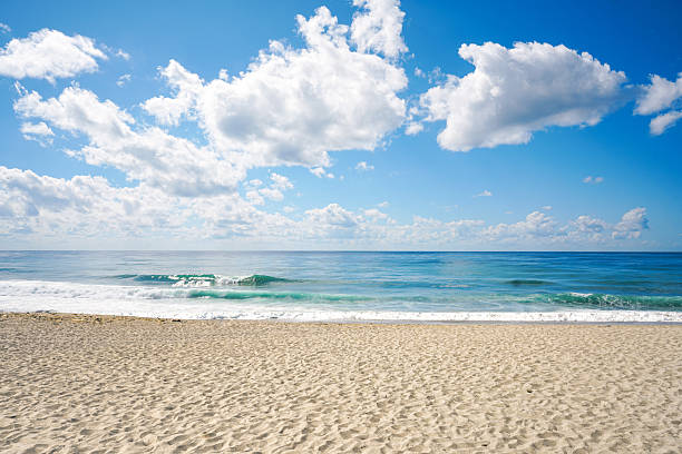Sunny sandy beach with turquoise waves under a blue sky and scattered white clouds. Peaceful summer seascape with wide horizon and natural light.