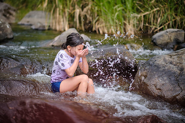 Little girl enjoying summer, splashing stream water with her hands while sitting on rocks in a flowing river outdoors