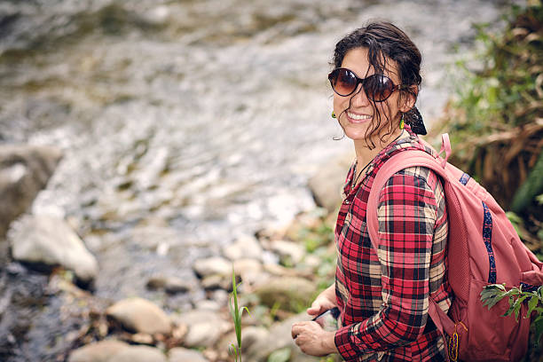 Happy woman hiking outdoors in nature, smiling while wearing sunglasses and a backpack by a flowing river