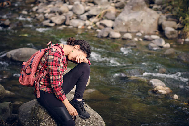 Young adult woman traveler sitting by a river, covering face with hand, experiencing sadness, despair, and emotional stress