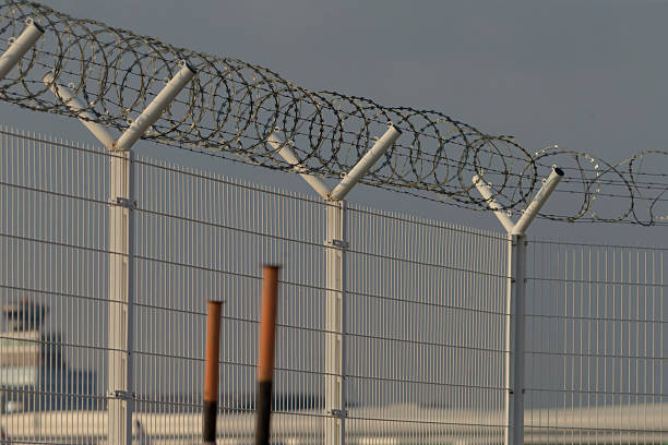 white runway boundary fence topped with continuous razor coils, grassy airfield beyond and distant tailfin visible, mood