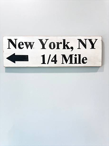 Vertical image of a New York directional road sign reading “New York, NY 1/4 Mile” with an arrow, mounted on a light colored wall. The minimal background, simple typography, and centered composition create a clean travel and navigation themed scene with ample copy space for text. This sign detail is ideal for illustrating road trips, directions, distance, and New York travel concepts in editorial and commercial projects.