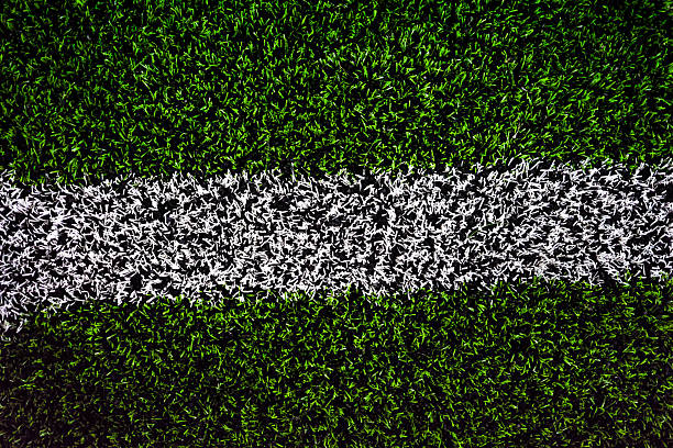 Close-up view of a white field marking line on green artificial turf. Detailed texture of synthetic grass used on football and sports fields, suitable as background, sports design, training, competition and copy space concepts.