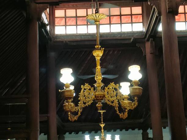 a classic brass chandelier suspended from the high wooden ceiling of a traditional Javanese mosque, illuminated by warm glowing lamps. The chandelier’s ornate floral and curved metal details reflect European-influenced lighting design adapted into local religious architecture. Behind it, tall dark wooden pillars, exposed ceiling beams, and clerestory windows emphasize the mosque’s spacious interior and natural ventilation system. The combination of soft artificial light, aged wood, and handcrafted ornamentation creates a calm, reverent atmosphere that highlights the historical and cultural character of the mosque, blending Islamic function with Javanese architectural heritage.
