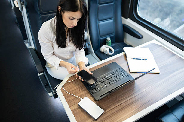 A woman intently charging her phone using a power bank while seated at a table on a train, illustrating how modern travel accommodates digital lifestyles and connectivity.
