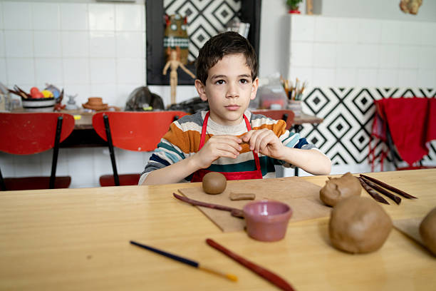 A young schoolgboy is wearing a red apron and sitting at a workbench while he is molding clay for a creative project during a clay workshop
