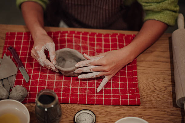 Hands forming raw clay into a bowl on a red checkered cloth, demonstrating handmade craft and creative expression