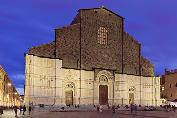 The Basilica di San Petronio is illuminated at night on Piazza Maggiore in Bologna, Emilia-Romagna, Italy. The illuminated church features its unfinished facade combining Gothic brickwork above with a pale marble lower section. The basilica is a prominent medieval religious building facing the city’s main public square.