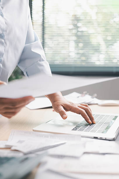 istock Office worker standing at desk while typing on laptop and reviewing multiple documents, illustrating business tasks, paperwork management, productivity and professional workflow in modern workplace. 2252296076