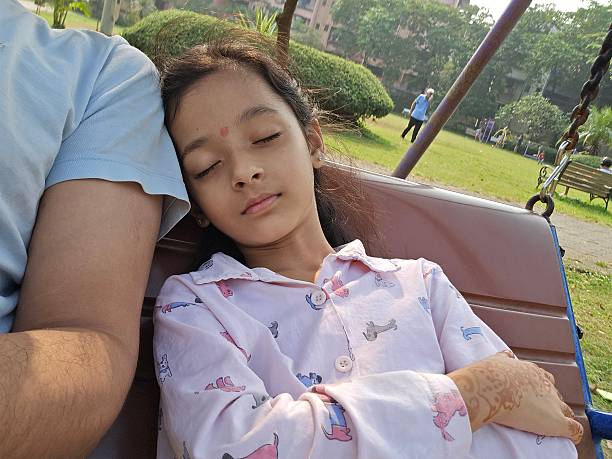 Close shot showing young girl resting on swing in peaceful park setting during daytime with soft light, relaxed expression, green trees, benches, and gentle outdoor atmosphere as she leans against adult beside her creating calm moment.