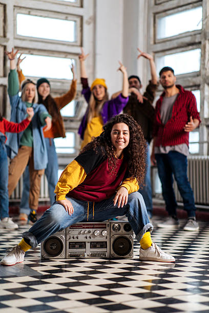 A diverse group of young people gather in a bright studio, with a central dancer beside a vintage boombox; Energetic, playful moment captures friendship, music, and urban dance vibes
