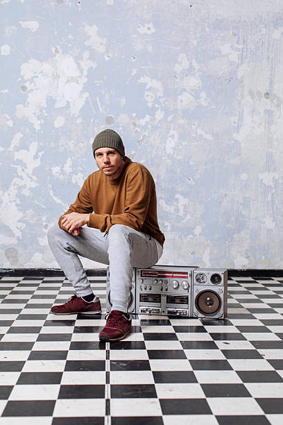 A casually dressed man sits on a retro boombox in a worn studio with a chipped blue wall and a black-and-white checkerboard floor, conveying urban, retro vibe and relaxed street style