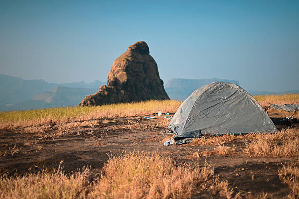 A camping tent set up on open grassland in a natural mountain landscape, surrounded by dry grass, scenic rock formations, and visible litter left on the ground. It represents outdoor travel, camping, and human impact on nature. The scene captures a camping site affected by waste and pollution during an outdoor journey, symbolizing environmental neglect, lack of responsible travel, and the contrast between natural beauty and littering. Ideal for concepts related to environmental awareness, pollution, responsible tourism, sustainable travel, eco travel education, conservation, outdoor lifestyle, camping ethics, and environmental protection.