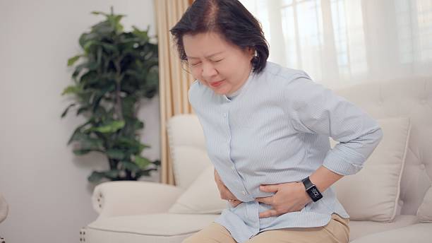 An elderly woman sits quietly inside her own home, using both hands to hold her abdomen possibly a health issue such as abdominal pain, indigestion, or severe cramps can occur without warning, as stomach pain and cramping through her body. Her expression shows a mixture of distress, fatigue, and vulnerability. Elderly Healthcare concept.