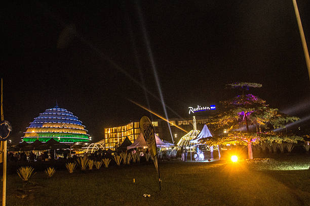 A stunning wide-angle color photograph capturing the modern skyline of Kigali at night. The image features the iconic Kigali Convention Centre dome glowing in national colors, the Radisson Blu hotel, and an illuminated monument under a canopy of decorative lights. The scene is filled with the energy of a nighttime event, featuring spotlights cutting through the sky, white event tents, and lush green landscaping. This image is a premier choice for themes of African urbanism, tourism, economic development, and modern infrastructure.