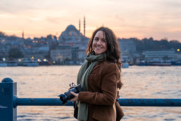 Woman holding a camera smiles while standing by the water with the city in the background