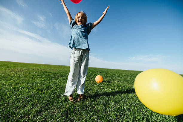 A cute little girl with her hair in twin buns diligently inflates a bright yellow balloon with a hand pump on a rooftop, with the cityscape blurred in the background, enjoying a playful day out.