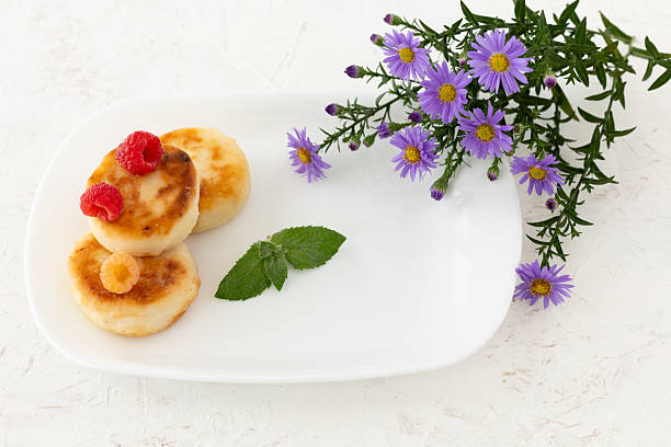 White porcelain plate with homemade curd fritters, raspberries and mint leaves, a bouquet of flowers on the white background. Top view.
