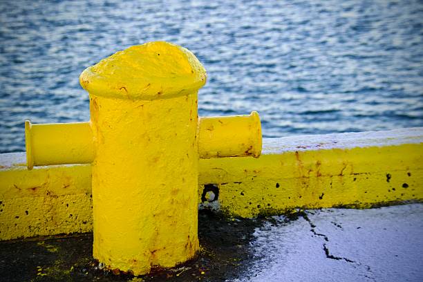 A close-up view of a weathered yellow harbour bollard set against blue ocean water along the seafront. The image captures industrial maritime infrastructure, winter conditions and strong colour contrast typical of Nordic coastal environments. Picture taken in Iceland.