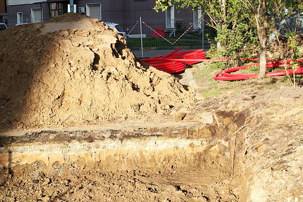 istock Open trench and large dirt pile showing different soil layers next to coiled red plastic pipes for utilities installation at a sunny urban construction site 2252332498
