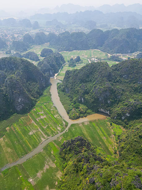 Drone shot of peaceful rivers snaking through limestone cliffs and emerald-green rice fields in Ninh Bình Province.