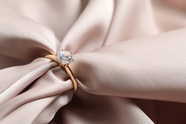istock Beautiful shiny gold engagement ring with large diamond gemstone on silk fabric background 2252346511