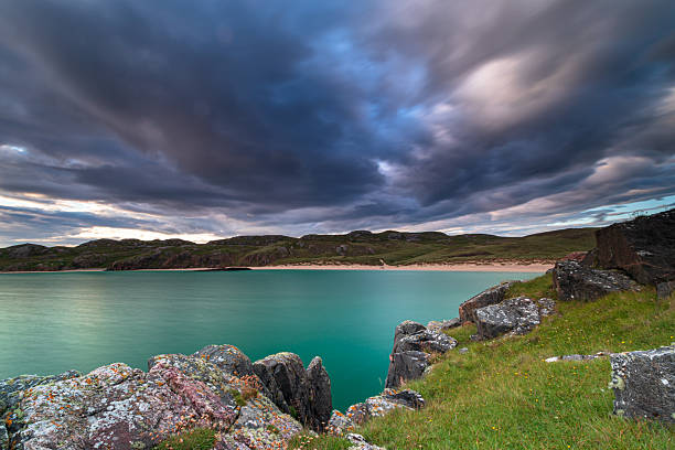 Oldshoremore beach - Turquoise water and rugged rocks frame a tranquil coastal scene. Dark storm clouds roll over grassy hills, creating a moody, dramatic mood that highlights nature, seascapes, and wide open space.